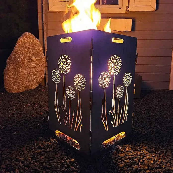 TiKo-Design Holzkohlegrill Feuerkorb-Pusteblumen - Durchmesser Grillfläche: 60 cm, Grau - Schwarze Metall-Feuerschale mit Blumenmotiven, beleuchtet von tanzenden Flammen im Freien.
