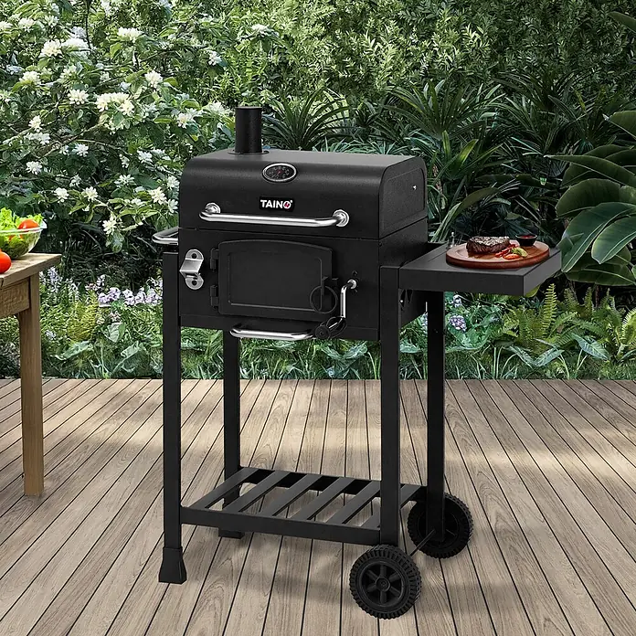 TAINO Holzkohlegrill HERO - Schwarz - Schwarzer Metall-Holzkohlegrill mit Rädern und Seitenablage, auf einer Holzterrasse im Garten.