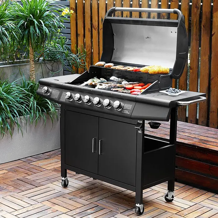 TAINO Gasgrill RED 6+1 -  - Ein schwarzer Gasgrill mit mehreren Brennern und darauf zubereitetem Essen auf einer Holzterrasse mit Pflanzen.