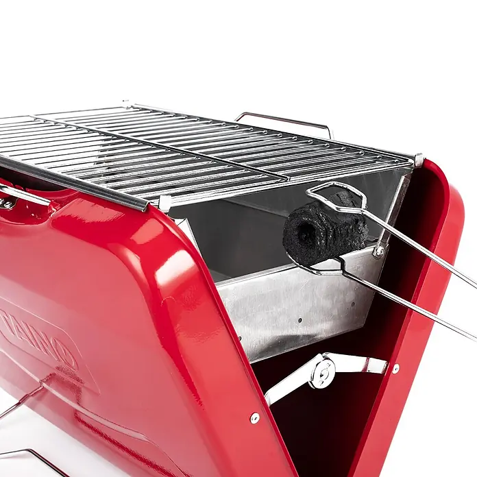 TAINO Holzkohlegrill MOX - Rot - Roter tragbarer Holzkohlegrill mit Edelstahlrost und Kohlebehälter. Eine Grillzange hält einen Brikett zum Anzünden bereit.
