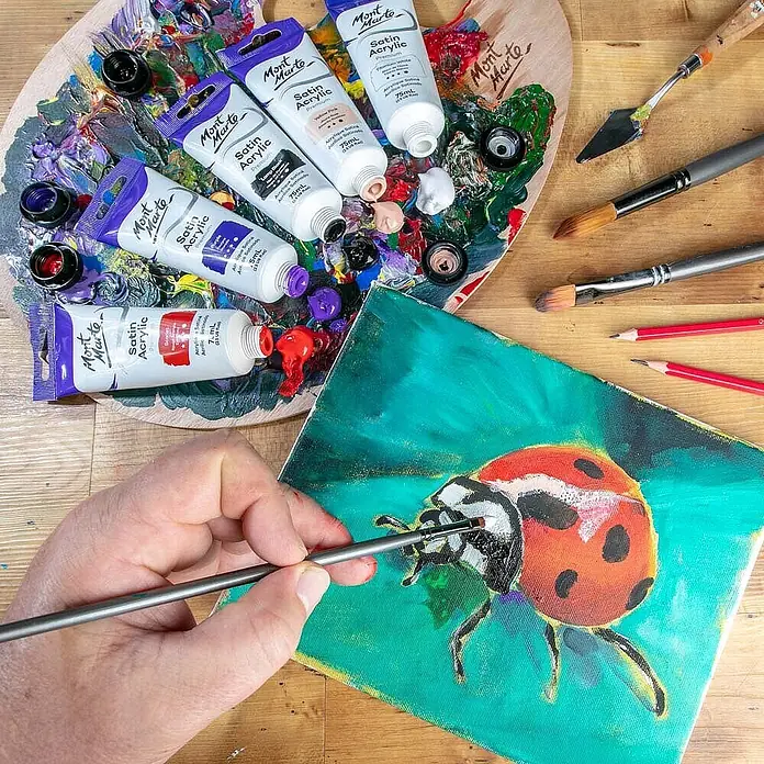 Mont Marte Acrylfarbe Premium - Titanweiß, 75 ml, Tube - Eine Hand malt einen Marienkäfer auf Leinwand mit Mont Marte Satin Acrylfarben, Pinseln und einer Holzpalette voller Farben.