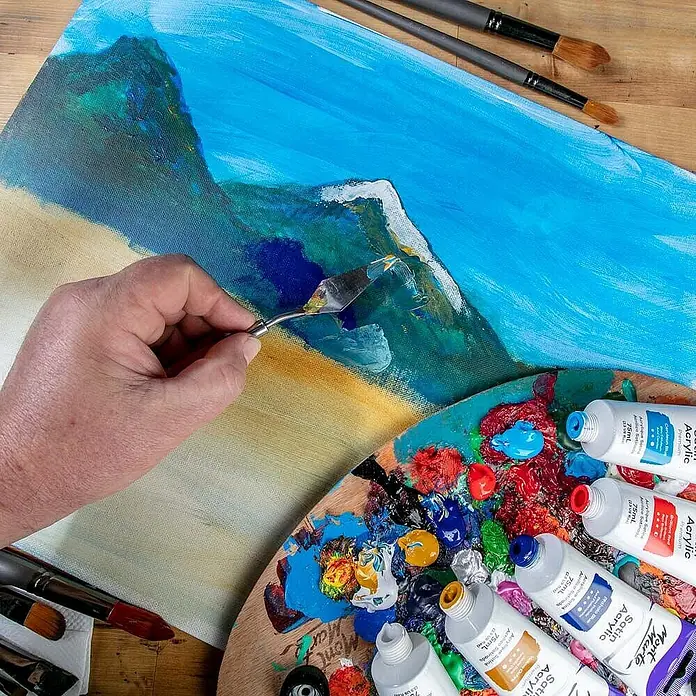 Mont Marte Acrylfarbe Premium - Ocker Gelb, 75 ml, Tube - Eine Hand verwendet einen Spachtel, um Farbe auf eine Leinwand aufzutragen, mit einer Palette voller bunter Acrylfarben und Tuben daneben.