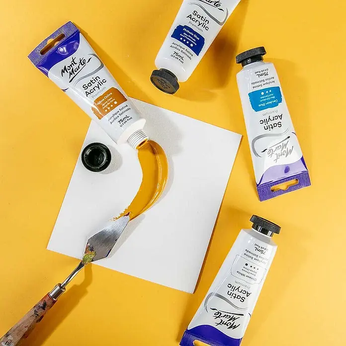 Mont Marte Acrylfarbe Premium - Ocker Gelb, 75 ml, Tube - Gelber Ocker Mont Marte Satin Acrylfarbe auf weißer Fläche, mit Spachtel verteilt, daneben blaue und weiße Farb-Tuben.