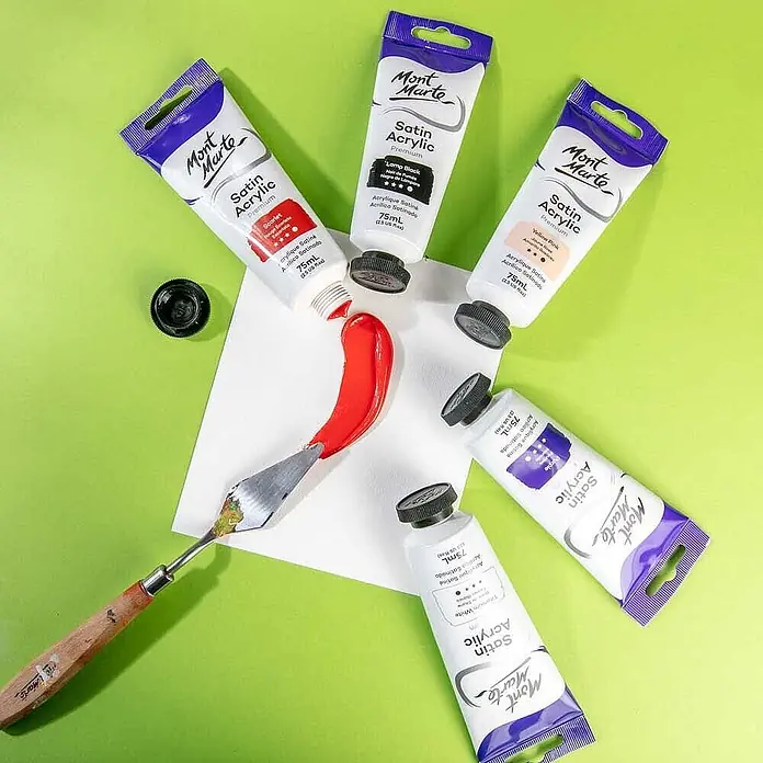 Mont Marte Acrylfarbe Premium - Scharlachrot, 75 ml, Tube - Mehrere Tuben Mont Marte Satin Acrylfarbe in verschiedenen Farben liegen um einen Spachtel mit roter Farbe auf einer weißen Fläche.