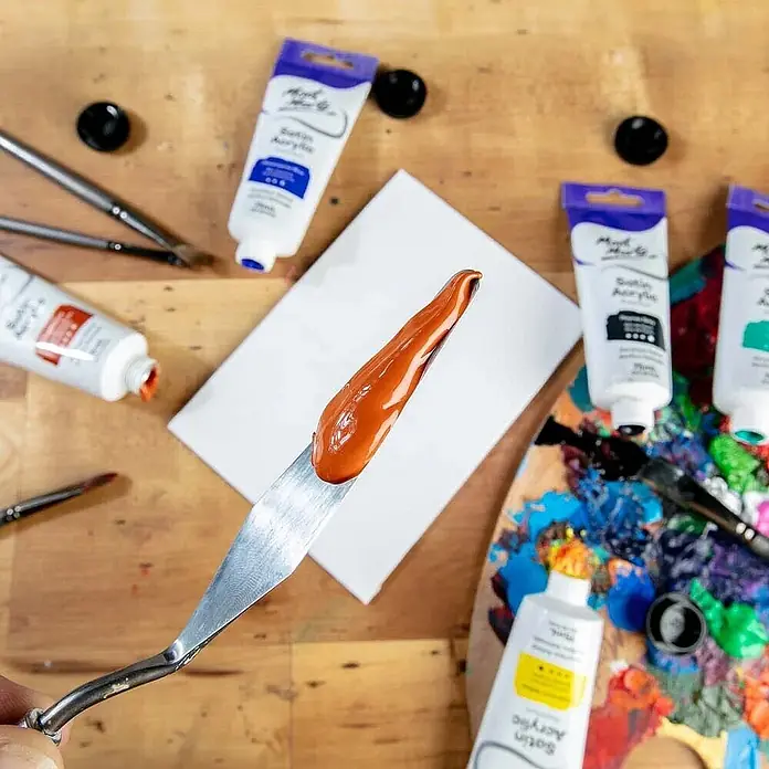 Mont Marte Acrylfarbe Premium - Gebrannte Siena, 75 ml, Tube - Orange Acrylfarbe auf einem Spachtel, umgeben von Satin-Acrylfarbentuben, Pinseln und einer farbenfrohen Künstlerpalette.