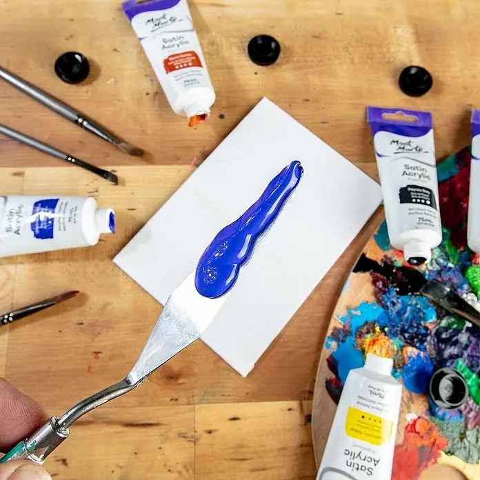 Mont Marte Acrylfarbe Premium - Ultramarinblau, 75 ml, Tube - Ein Metallspachtel mit leuchtend blauer Acrylfarbe, über einer kleinen weißen Leinwand. Kunstzubehör umgibt es.