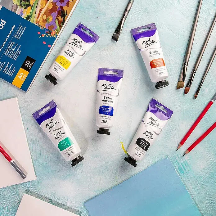 Mont Marte Acrylfarbe Premium - Ultramarinblau, 75 ml, Tube - Fünf Mont Marte Satin Acrylfarbtuben (75ml) in verschiedenen Farben, umgeben von Pinseln, Skizzenblock und Bleistiften.