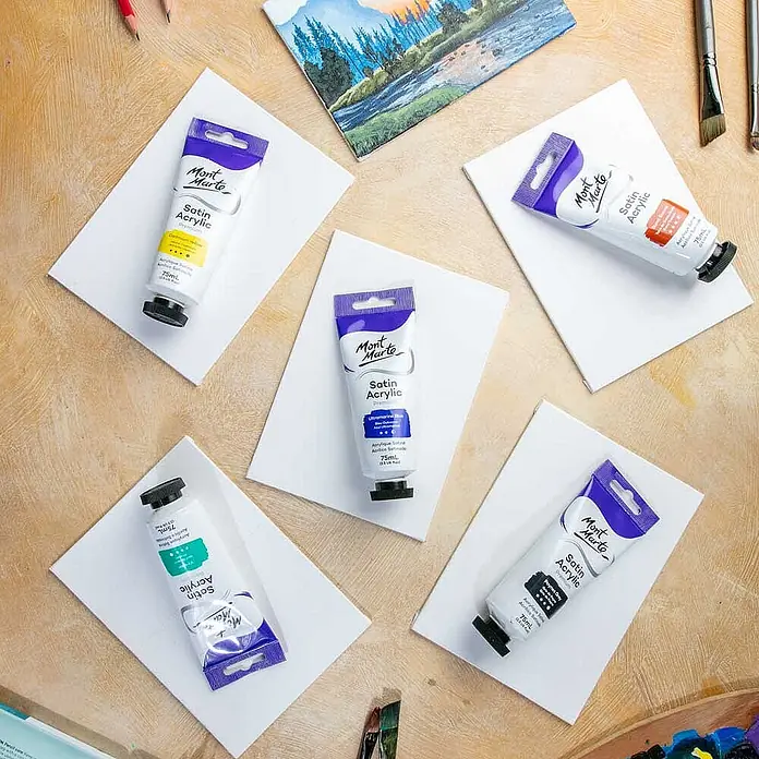 Mont Marte Acrylfarbe Premium - Ultramarinblau, 75 ml, Tube - Mont Marte Satin Acrylfarben in verschiedenen Farben auf weißen Leinwänden, umgeben von Kunstzubehör.