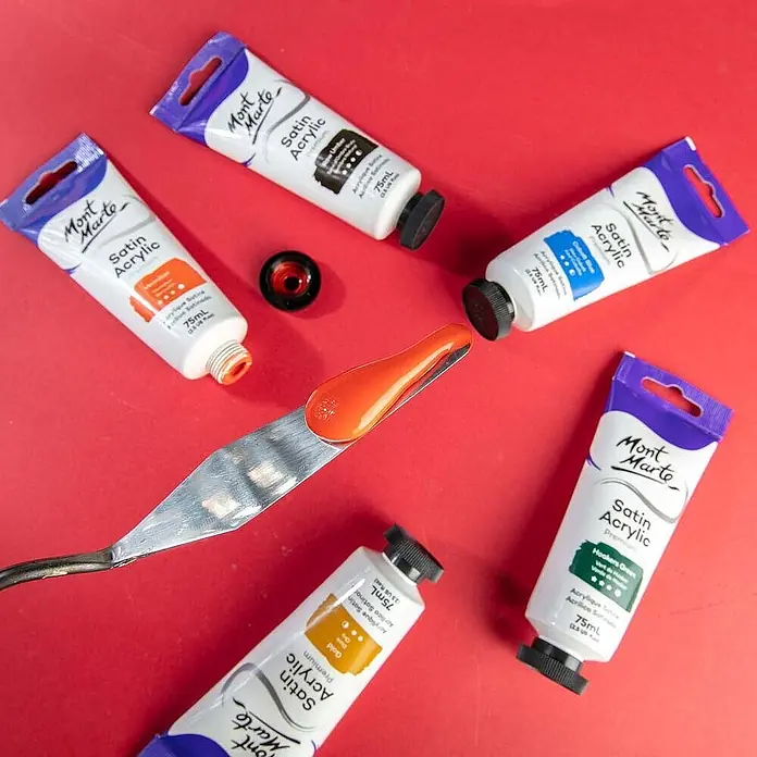 Mont Marte Acrylfarbe Premium - Zinnoberrot, 75 ml, Tube - Mont Marte Satin Acrylfarben-Tuben in verschiedenen Farben, mit orangefarbener Farbe auf einem Spachtel, Künstlerbedarf auf Rot.