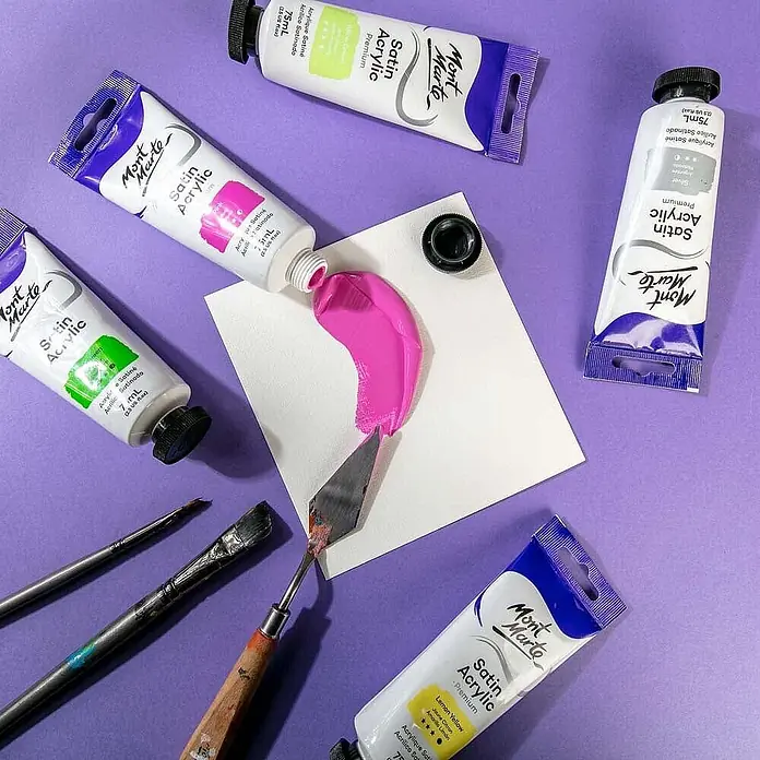 Mont Marte Acrylfarbe Premium - Pink, 75 ml, Tube - Mont Marte Satin Acrylfarbtuben, eine pinke Farbe auf eine Karte drückend, mit Spachtel und weiteren Künstlermaterialien.