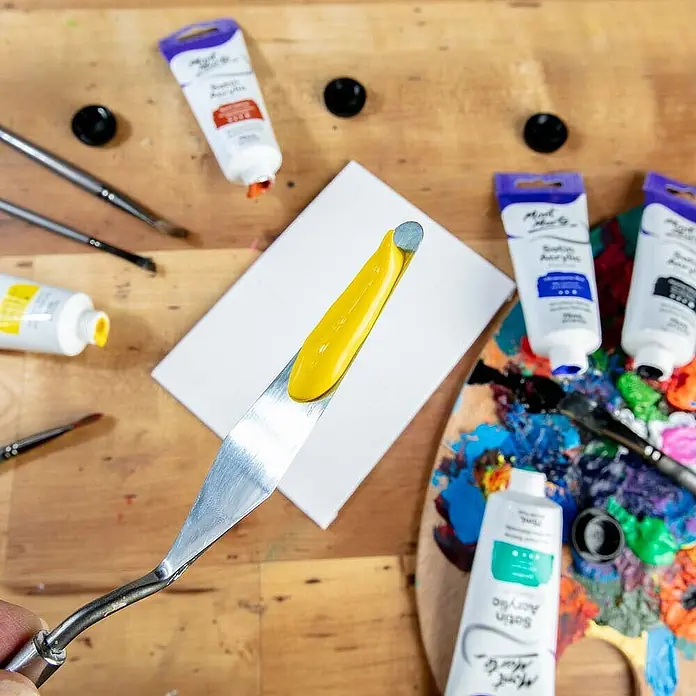 Mont Marte Acrylfarbe Premium - Kadmiumgelb, 75 ml, Tube - Ein Metall-Malmesser hält einen Klecks hellgelber Farbe, auf einer kleinen weißen Leinwand inmitten anderer Kunstmaterialien auf einem Holztisch.