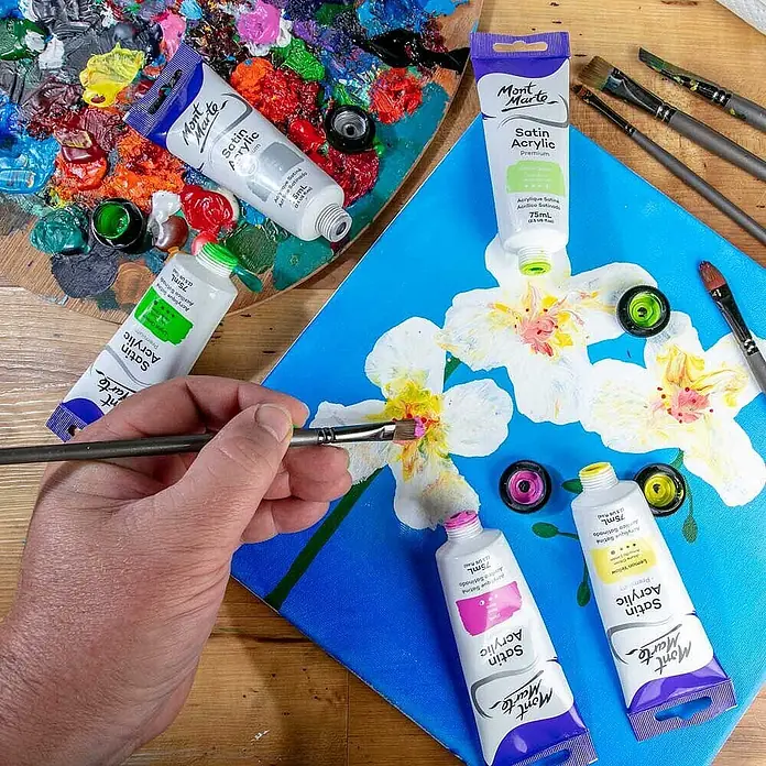 Mont Marte Acrylfarbe Premium - Pink, 75 ml, Tube - Mont Marte Satin Acrylfarben in verschiedenen Farben, verwendet, um weiße Blumen auf eine blaue Leinwand zu malen. Eine Hand malt mit einem Pinsel.