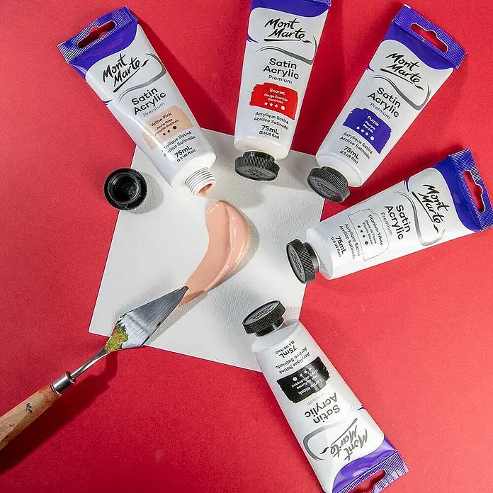 Mont Marte Acrylfarbe Premium - Gelbrosa, 75 ml, Tube - Mehrere Mont Marte Satin Acrylfarbtuben um einen Spachtel, der pfirsichfarbene Farbe auf weißer Oberfläche mischt.