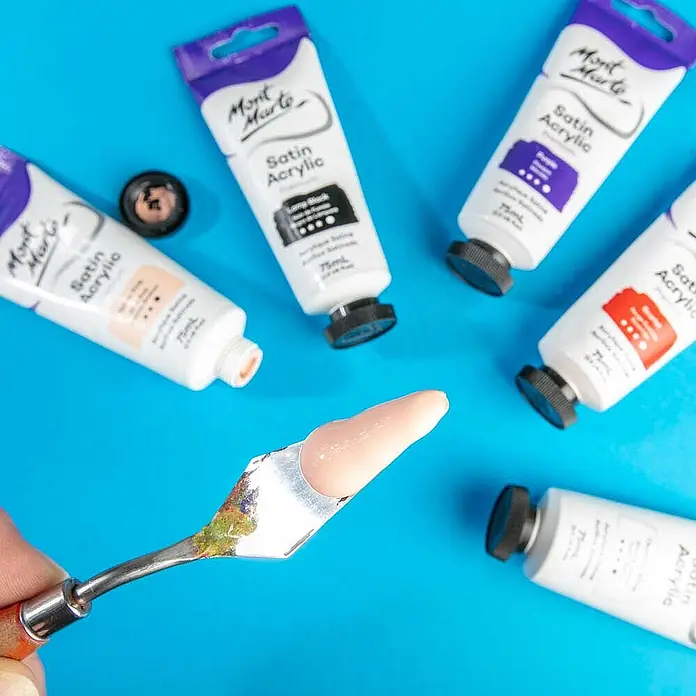 Mont Marte Acrylfarbe Premium - Gelbrosa, 75 ml, Tube - Mont Marte Satin Acrylfarben-Tuben mit beigefarbener Farbe auf Spachtel, auf blauem Untergrund.