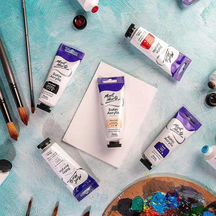 Mont Marte Acrylfarbe Premium - Gelbrosa, 75 ml, Tube - Mont Marte Satin Acrylfarben-Tuben, Pinsel und eine Palette mit bunten Farben liegen auf blauem Untergrund, bereit zum Malen.