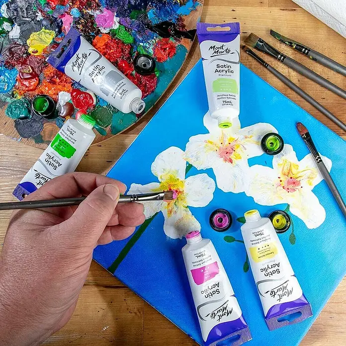 Mont Marte Acrylfarbe Premium - Hellgrün, 75 ml, Tube - Mont Marte Satin Acrylfarben in Tuben, von einer Hand zum Malen lebhafter Blumen auf blauer Leinwand mit Pinseln verwendet.