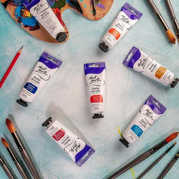 Mont Marte Acrylfarbe Premium - Brillantrot, 75 ml, Tube - Mehrere Mont Marte Satin Acrylfarben-Tuben in verschiedenen Farben, mit Pinseln und Palette, auf blauem Untergrund.