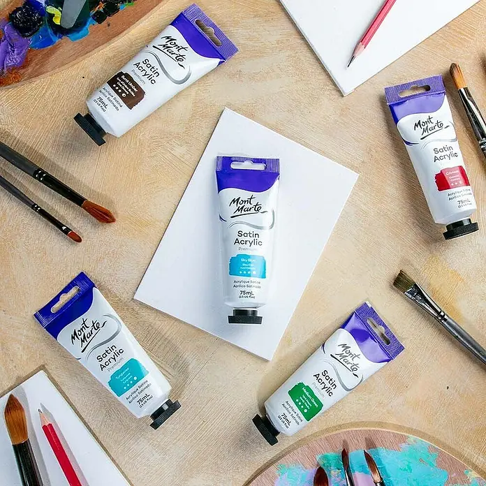 Mont Marte Acrylfarbe Premium - Himmelblau, 75 ml, Tube - Tuben Mont Marte Satin Acrylfarbe in verschiedenen Farben, Pinsel und Palette auf Holzoberfläche arrangiert.
