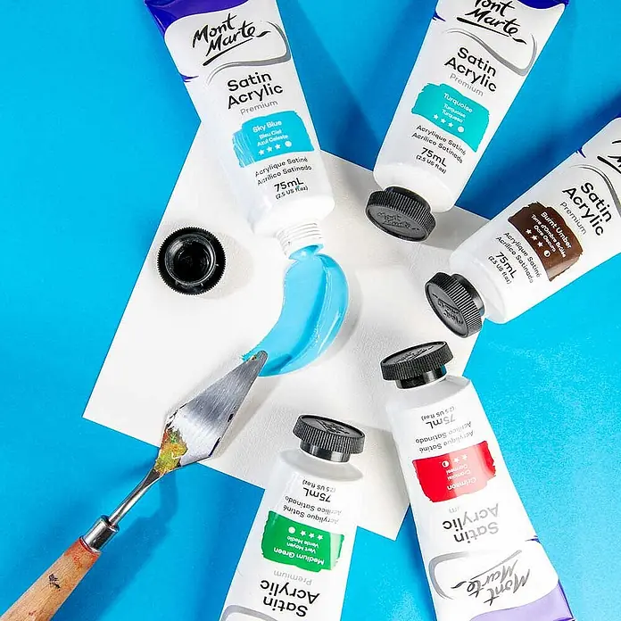 Mont Marte Acrylfarbe Premium - Himmelblau, 75 ml, Tube - Mont Marte Satin Acrylfarbtuben in verschiedenen Farben, eine Tube gibt hellblaue Farbe auf eine weiße Oberfläche neben einem Spachtel ab.