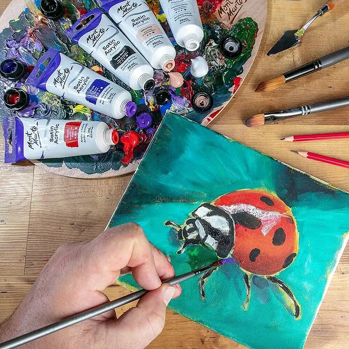 Mont Marte Acrylfarbe Premium - Violett, 75 ml, Tube - Mont Marte Satin Acrylfarben-Tuben auf einer Palette, von einer Hand zum Bemalen eines Marienkäfers auf Leinwand verwendet.