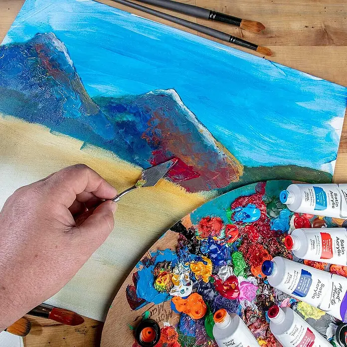 Mont Marte Acrylfarbe Premium - Rosenrot, 75 ml, Tube - Hand trägt mit Spachtel dicke rote Acrylfarbe auf eine Leinwand mit Bergmotiv auf, umgeben von Palette und Farbtuben.