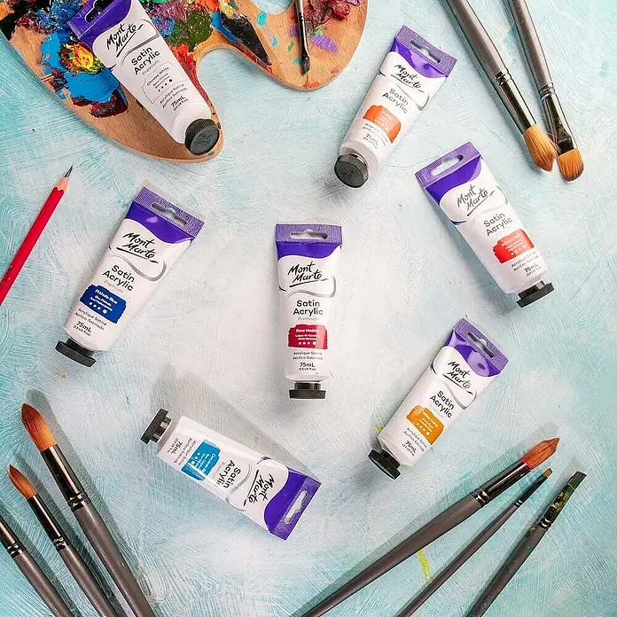 Mont Marte Acrylfarbe Premium - Rosenrot, 75 ml, Tube - Mehrere Tuben Mont Marte Satin Acrylfarben in verschiedenen Farben, Pinsel und eine Künstlerpalette auf einem strukturierten blauen Untergrund.