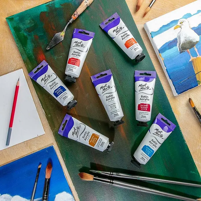 Mont Marte Acrylfarbe Premium - Chinesisch Weiß, 75 ml, Tube - Mehrere Tuben Mont Marte Satin Acrylfarbe in verschiedenen Farben, umgeben von Pinseln und bemalten Leinwänden.