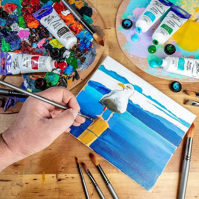 Mont Marte Acrylfarbe Premium - Gebrannte Umbra, 75 ml, Tube - Eine Person malt mit Mont Marte Satin Acrylfarben und Pinseln ein Möwenbild auf Leinwand. Bunte Farbtuben und Paletten liegen bereit.