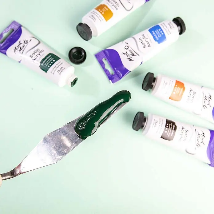 Mont Marte Acrylfarbe Premium - Hookers Grün, 75 ml, Tube - Mehrere Mont Marte Satin Acrylfarben-Tuben, mit einem Spachtel, der einen Klecks dunkelgrüner Farbe hält.