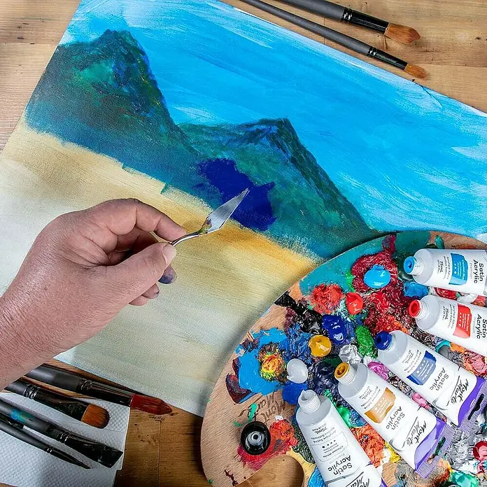 Mont Marte Acrylfarbe Premium - Phthaloblau, 75 ml, Tube - Eine Hand trägt mit einem Malmesser Mont Marte Satin Acrylfarbe auf eine Landschaftsleinwand auf, umgeben von Farbtuben und Pinseln.