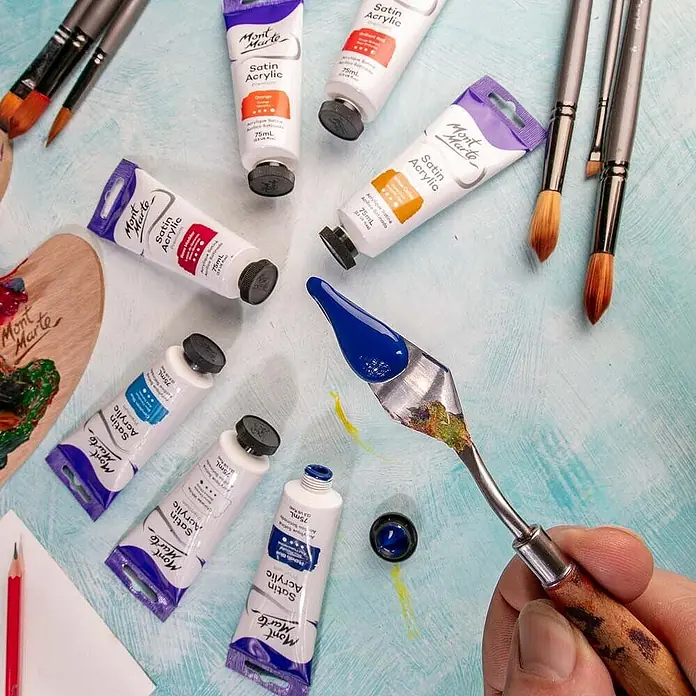 Mont Marte Acrylfarbe Premium - Phthaloblau, 75 ml, Tube - Eine Auswahl an Mont Marte Satin Acrylfarben-Tuben in verschiedenen Farben, Künstlerpinsel und ein Spachtel mit blauer Farbe.