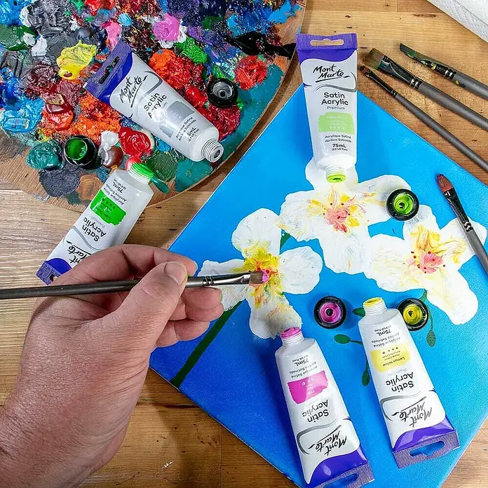 Mont Marte Acrylfarbe Premium - Zitronengelb, 75 ml, Tube - Mont Marte Satin Acrylfarben in Tuben auf Holz, eine Tube auf blauer Leinwand mit gemalten weißen Blumen, Pinsel und Palette.