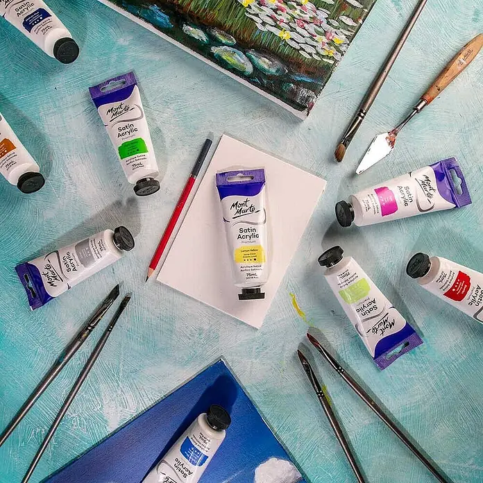 Mont Marte Acrylfarbe Premium - Zitronengelb, 75 ml, Tube - Mont Marte Satin Acrylfarben, Pinsel und Kunstzubehör auf blauem Untergrund mit einer teilweise bemalten Leinwand.