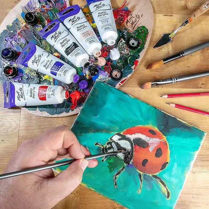 Mont Marte Acrylfarbe Premium - Lampenschwarz, 75 ml, Tube - Künstlerhand malt mit Mont Marte Satin Acrylfarben aus Tuben auf einer Palette einen roten Marienkäfer auf grüne Leinwand, umgeben von Pinseln.