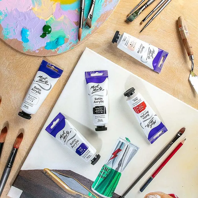 Mont Marte Acrylfarbe Premium - Lampenschwarz, 75 ml, Tube - Mont Marte Satin Acrylfarben-Tuben, Pinsel und eine Palette mit gemischten Farben auf einem Künstlerarbeitsplatz, neben einer unfertigen Leinwand.