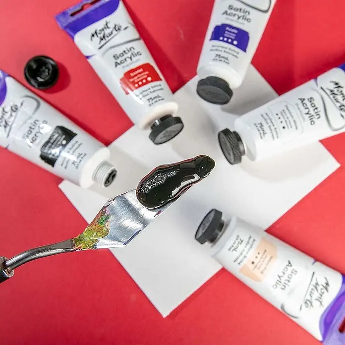 Mont Marte Acrylfarbe Premium - Lampenschwarz, 75 ml, Tube - Mont Marte Satin Acrylfarben-Tuben umgeben einen Spachtel mit einem Klecks schwarzer Farbe.