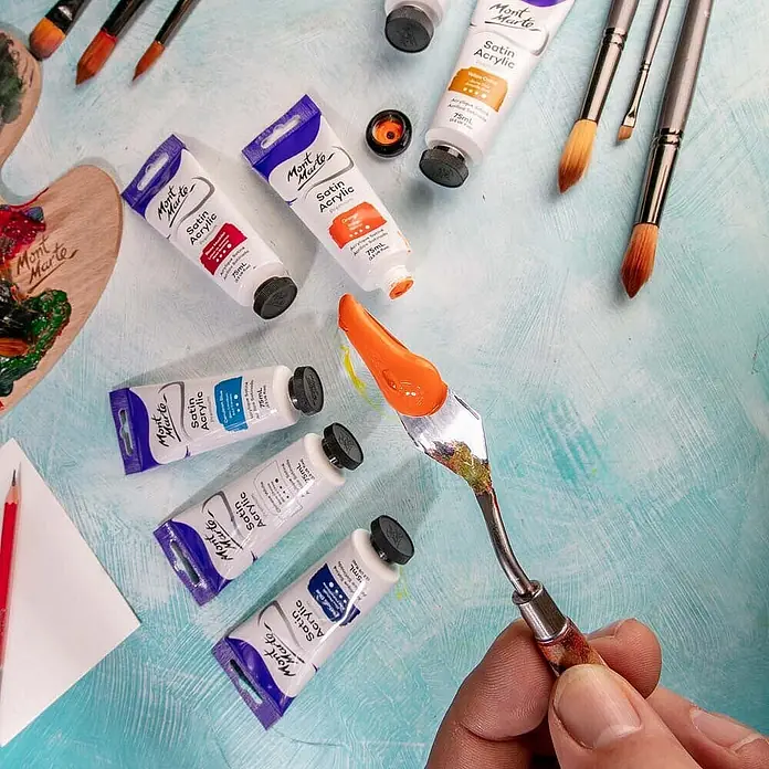 Mont Marte Acrylfarbe Premium - Orange, 75 ml, Tube - Mont Marte Satin Acrylfarben in Tuben, eine Hand hält ein Malmesser mit orangefarbener Farbe, Pinsel und Palette, bereit für Kunst.