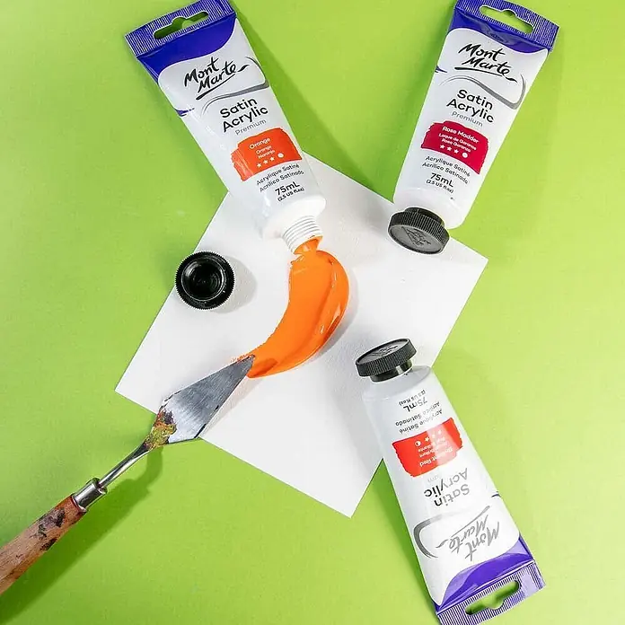 Mont Marte Acrylfarbe Premium - Orange, 75 ml, Tube - Drei Mont Marte Satin Acrylfarbtuben (orange, rosenrot, rot), wobei orange Farbe mit einem Spachtel auf eine Fläche gedrückt wird.