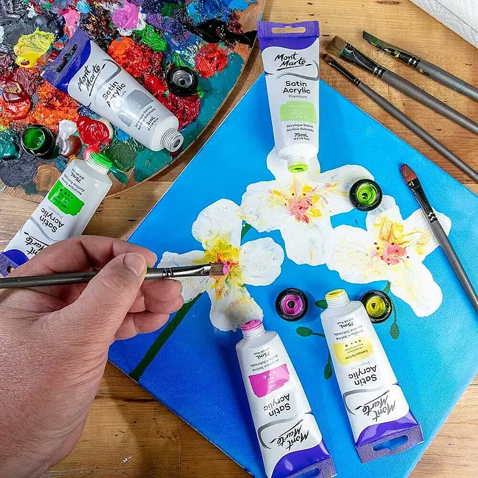 Mont Marte Acrylfarbe Premium - Gelbgrün, 75 ml, Tube - Künstlerhand malt weiße Blumen mit rosa und gelben Mont Marte Satin Acrylfarben auf blauer Leinwand, umgeben von Tuben und Pinseln.