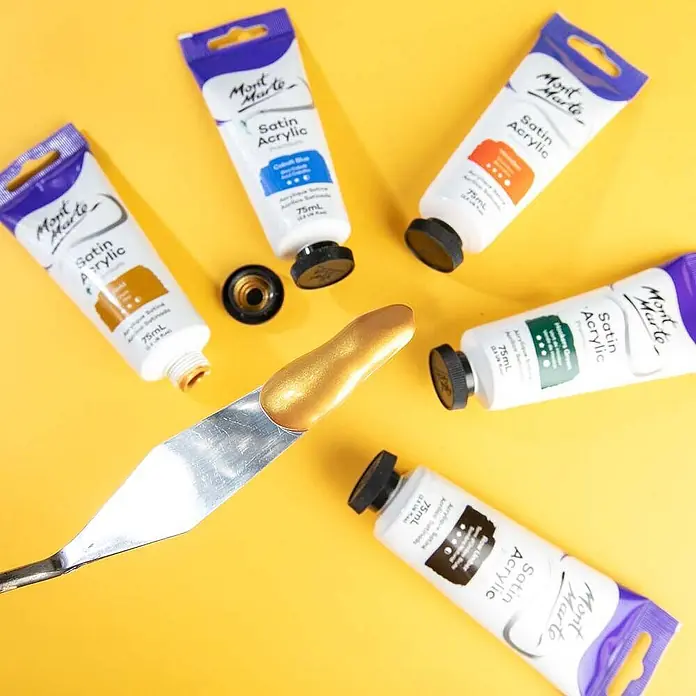 Mont Marte Acrylfarbe Premium - Gold, 75 ml, Tube - Mont Marte Satin Acrylfarben-Tuben und ein Spachtel mit goldener Farbe auf gelbem Untergrund.