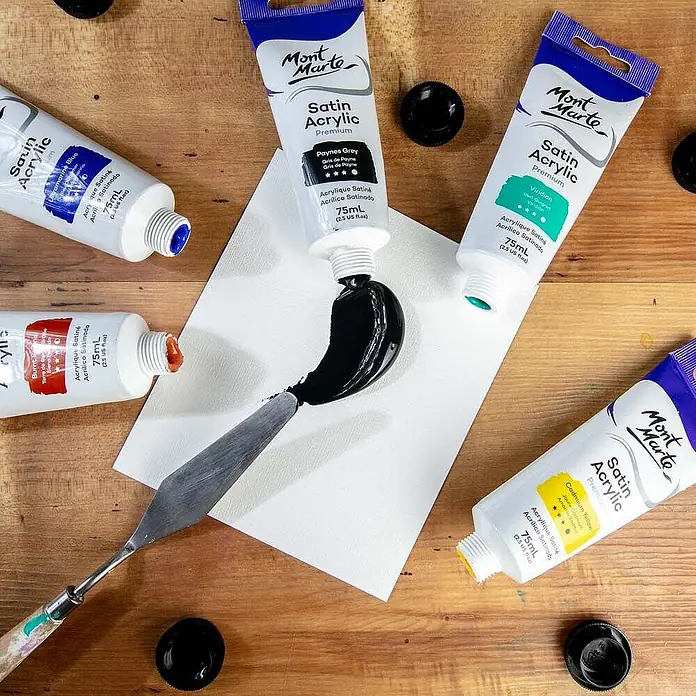 Mont Marte Acrylfarbe Premium - Paynesgrau, 75 ml, Tube - Mont Marte Satin Acrylfarbtuben in verschiedenen Farben, ein Metallspachtel und schwarze Farbe auf weißem Papier auf Holzoberfläche.
