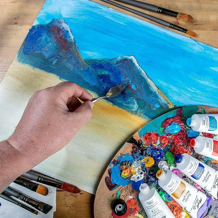 Mont Marte Acrylfarbe Premium - Zeruleanblau, 75 ml, Tube - Eine Künstlerhand malt mit einem Spachtel eine Landschaft auf Leinwand, umgeben von einer Holzpalette mit Acrylfarben und Pinseln.
