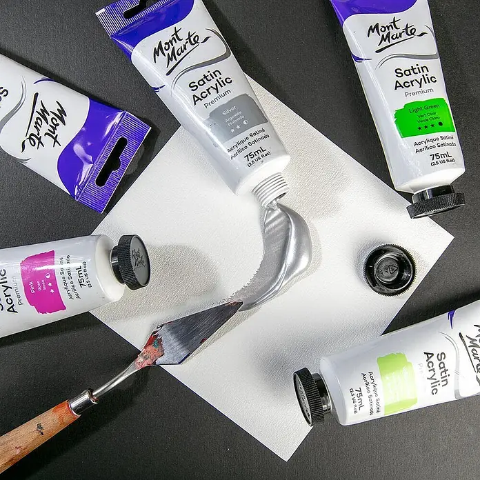 Mont Marte Acrylfarbe Premium - Silber, 75 ml, Tube - Mehrere Mont Marte Satin Acryl Premium Farbtuben (Silber, Pink, Hellgrün), mit Silberfarbe auf eine Oberfläche gedrückt neben einem Spachtel.