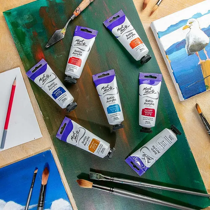 Mont Marte Acrylfarbe Premium - Zeruleanblau, 75 ml, Tube - Mehrere 75ml Mont Marte Satin Acrylfarben-Tuben inmitten von Pinseln, einem Spachtel und bemalten Leinwänden.