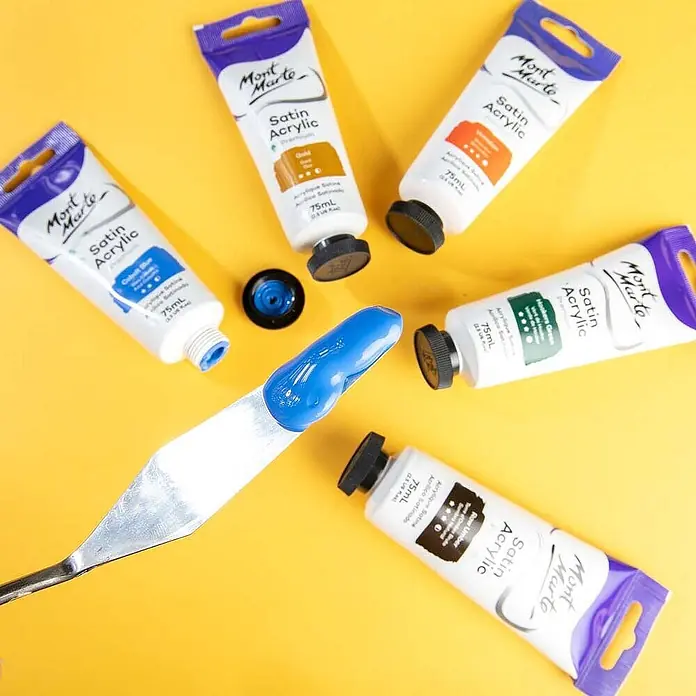 Mont Marte Acrylfarbe Premium - Kobaltblau, 75 ml, Tube - Verschiedene Mont Marte Satin Acrylfarben-Tuben und ein Spachtel mit blauer Farbe auf gelbem Untergrund.