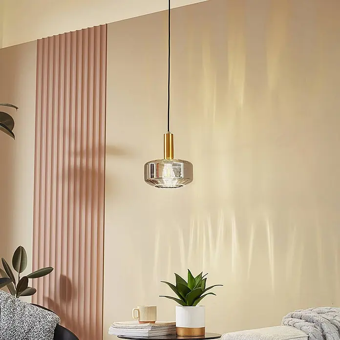 Lindby Pendelleuchte Hildur - alu / grau / zink, rauchgrau, messing, E27 - Eine schlichte Pendelleuchte mit klarem Glasschirm und Goldakzent hängt an einem schwarzen Kabel und beleuchtet einen modernen Wohnraum.