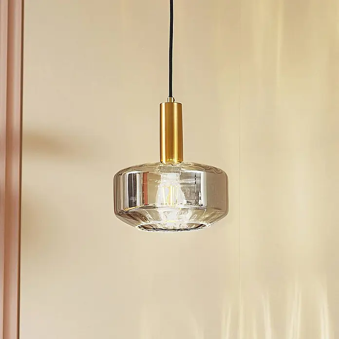 Lindby Pendelleuchte Hildur - alu / grau / zink, rauchgrau, messing, E27 - Pendelleuchte aus Gold und Rauchglas mit sichtbarer Glühbirne, an einer hellen Wand hängend.