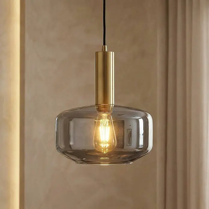 Lindby Pendelleuchte Hildur - alu / grau / zink, rauchgrau, messing, E27 - Eine moderne Pendelleuchte mit Messingfassung und Rauchglas-Schirm, einer beleuchteten Edison-Birne, hängt in einem Raum.