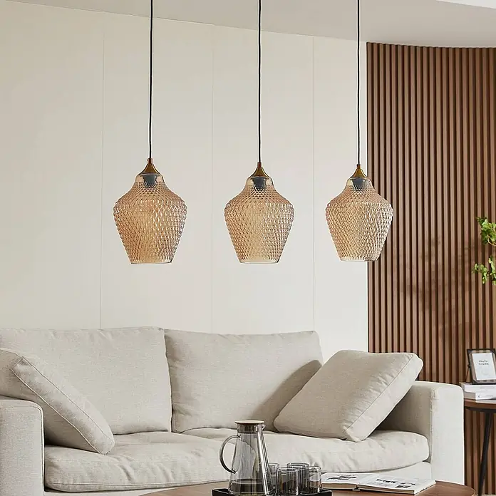 Lindby Pendelleuchte Drakar - L x B x H: 97 x 22 x 32 cm, creme / amber, amber, gold, E27 - Drei Pendelleuchten aus bernsteinfarbenem Strukturglas mit Messingakzenten hängen über einem Sofa im Wohnzimmer.