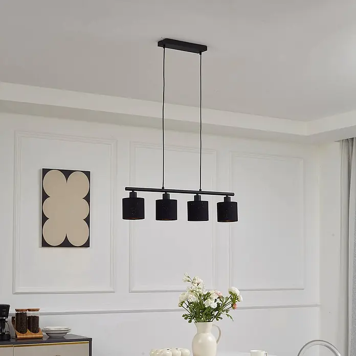 Lindby Pendelleuchte Vasilia - L x B x H: 75 x 12 x 11 cm, schwarz, Schwarz, gold, E14 - Schwarzer linearer Kronleuchter mit vier zylindrischen Schirmen, hängt über einem Esstisch in einem modernen Raum.